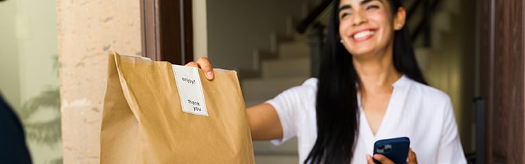 Closeup of a joyful latin woman accepting a food delivery in a brown paper bag at her doorstep