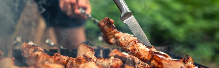 Person preparing  barbecued delicious grilled meats with smoke on fired charcoals. Hot BBQ closeup with nature on background