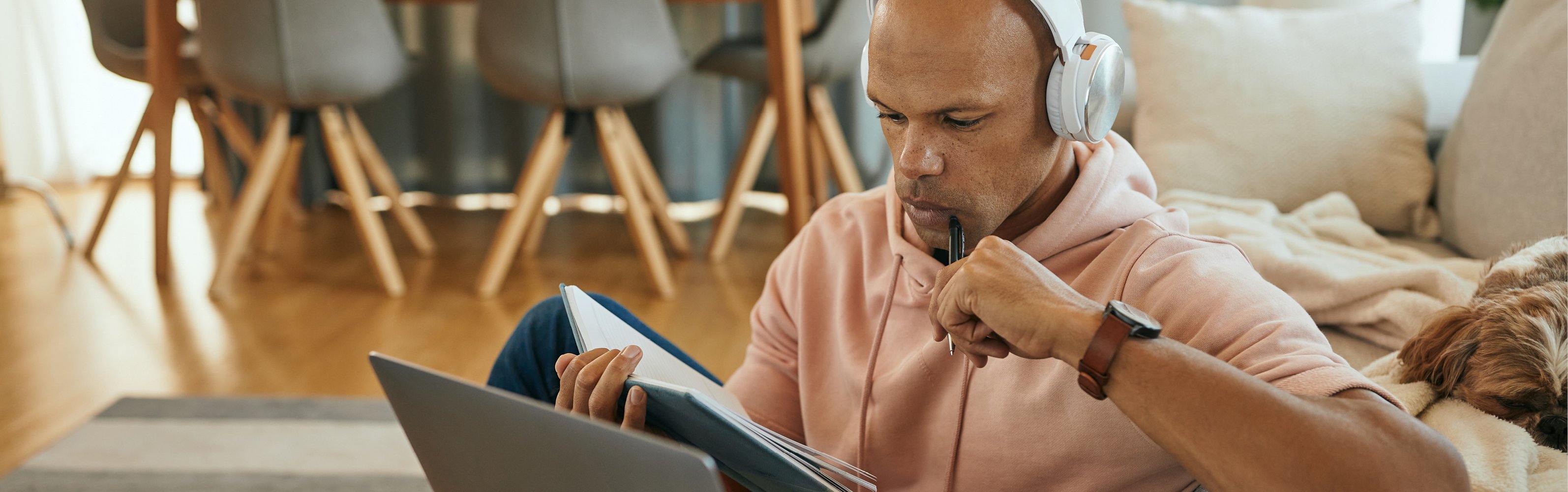 Young black man with headphones using laptop and reading notes while studying at home