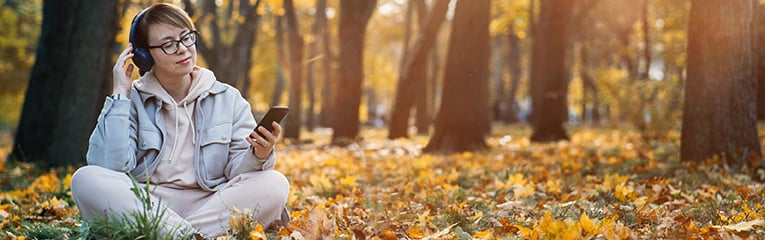 Caucasian middle aged woman in earphones listening to music, meditation app on smartphone and meditating in lotus pose at autumn park.