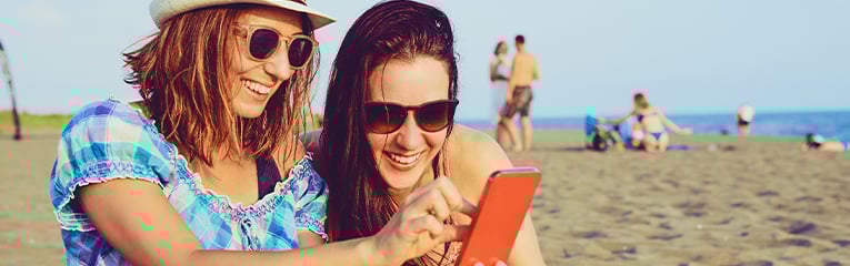 Girl friends on beach looking at cell phone on vacation taking a selfie.