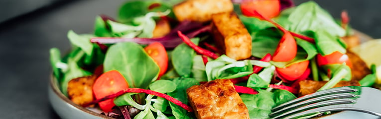 Healthy salad with roasted tempeh, fresh cherry tomatos, beetroot straws, spinash and lettuce leaves on plate.