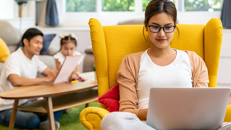 Young woman sitting on a yellow recliner on laptop with mother and sister reading in background.