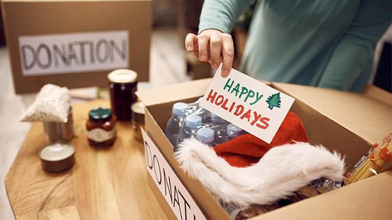 Close-up of woman donating food for charity and adding greeting card with 'Happy Holidays' message into a box.