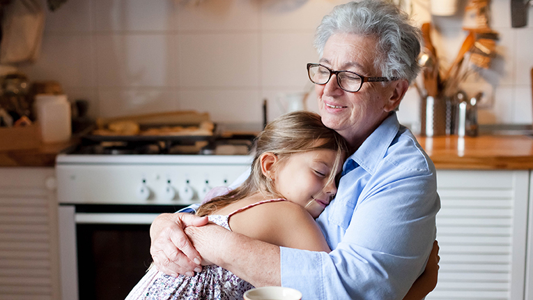 Smiling Causian grandmother sitting down hugging her granddaughter in kitchen