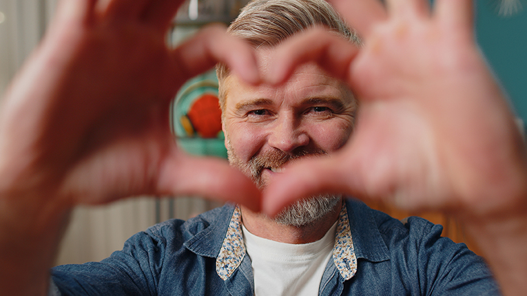 Happy middle-aged senior old man at home couch makes symbol of love, showing heart sign to camera