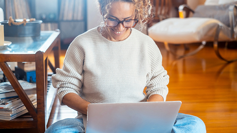 One adult woman working on laptop alone at home sitting on the wooden floor.