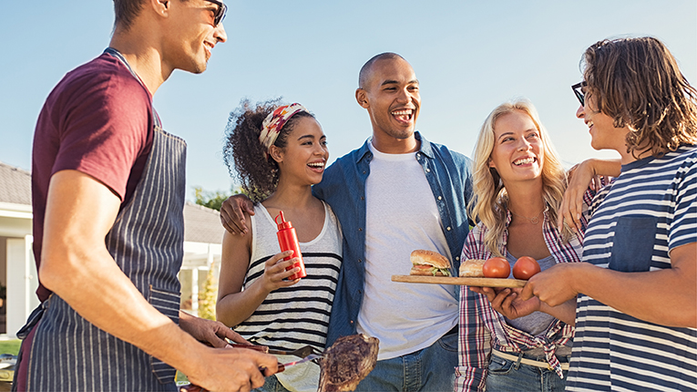 Young multiethnic friends having fun grilling meat enjoying bbq party and eating dinner outdoors. Men and women standing around grilling and eating. 