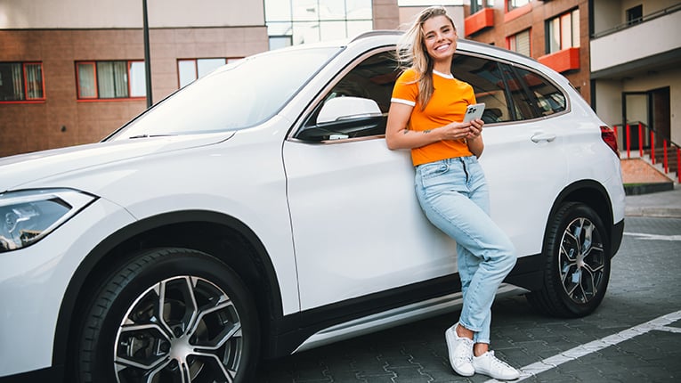 Young woman leaning against a white car outdoors in an urban area holding a smartphone and smiling joyfully on a sunny day stock photo