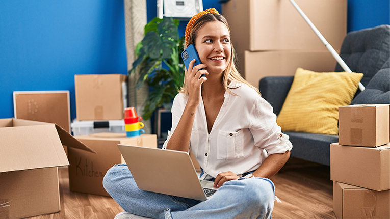 Young woman using laptop and talking on smartphone at new home