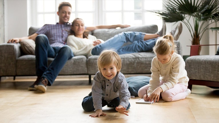 Happy family with cute kids playing while parents relax on sofa at home together, smiling active boy entertaining with toy car near his sister on the floor.  