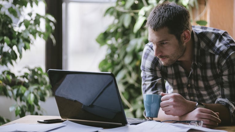 Man-searching-for-the-best-personal-loan-for-debt-consolidation-on-his-laptop-while-drinking-a-cup-of-coffee-and-reviewing-his-bills.