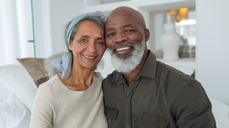 Front view of diverse senior couple smiling while sitting on couch inside a room in beach house.