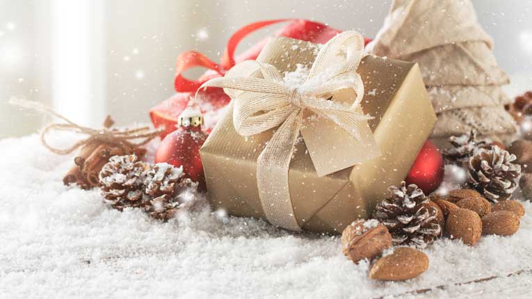 Gift box wrapped in gold wrapping paper with surrounded by pine cones sitting on fake snow. 